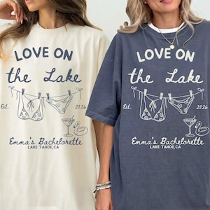 May include: Two short-sleeve crewneck t-shirts, one ivory and one denim blue, featuring a graphic print with the text LOVE ON the Lake, Est. 2026, Emma's Bachelorette, LAKE TAHOE, CA, a clothesline with two bikinis, a cocktail, and a flamingo.