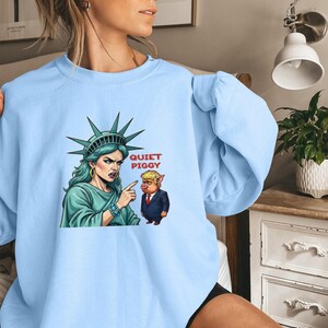 May include: Light blue sweatshirt featuring a graphic of the Statue of Liberty pointing at a cartoon pig in a suit. The text "QUIET PIGGY" is above the pig. The Statue of Liberty is wearing a crown and has green hair.