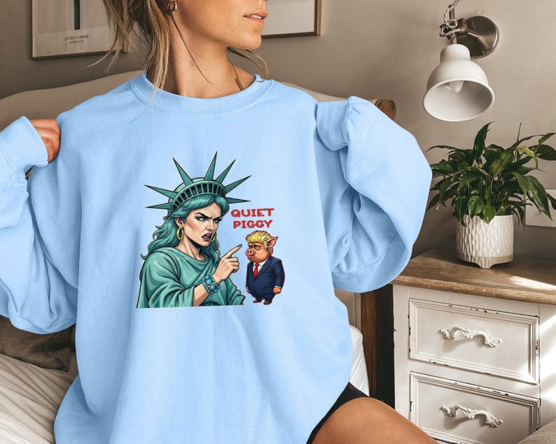 May include: Light blue sweatshirt featuring a graphic of the Statue of Liberty pointing at a cartoon pig in a suit. The text "QUIET PIGGY" is above the pig. The Statue of Liberty is wearing a crown and has green hair.