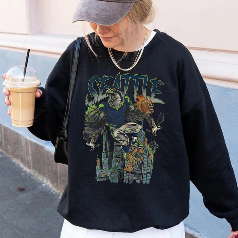 May include: Black sweatshirt with a graphic featuring the word "SEATTLE" in green and blue, along with an eagle-like figure and city skyline. The design incorporates shades of green, blue, and orange. The sweatshirt is worn by a person.
