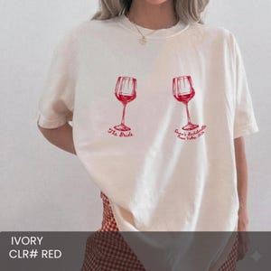 May include: An ivory short-sleeve t-shirt featuring two red wine glass graphics. The left glass is labeled The Bride, and the right glass features the text Emma's Bachelorette Napa Valley 2026. The shirt has a crew neckline and a relaxed fit.