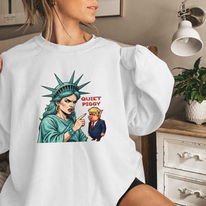 May include: White sweatshirt featuring a graphic of the Statue of Liberty pointing at a pig in a suit, with the text "QUIET PIGGY" in red. The Statue of Liberty is depicted with a stern expression.