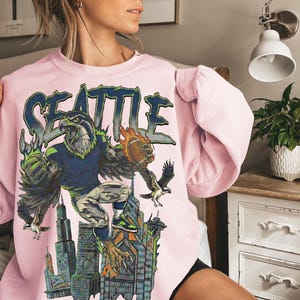 May include: A light pink sweatshirt featuring a graphic of a football-playing hawk with the word "SEATTLE" in blue and green. The hawk is set against a cityscape with a football. The sweatshirt has long sleeves.