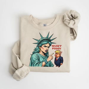 May include: Beige sweatshirt featuring a graphic of the Statue of Liberty pointing at a pig in a suit, with the text "Quiet Piggy". The Statue of Liberty is depicted with teal hair and a teal dress.