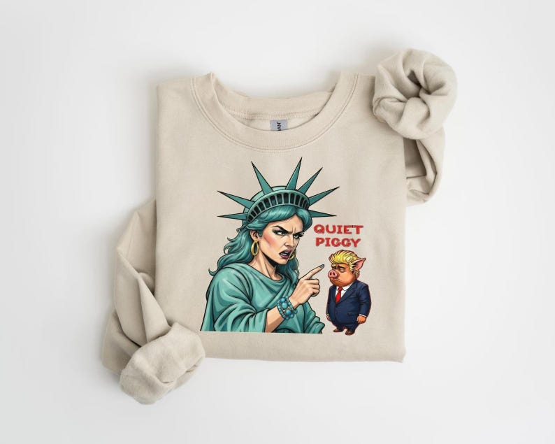 May include: Beige sweatshirt featuring a graphic of the Statue of Liberty pointing at a pig in a suit, with the text "Quiet Piggy". The Statue of Liberty is depicted with teal hair and a teal dress.