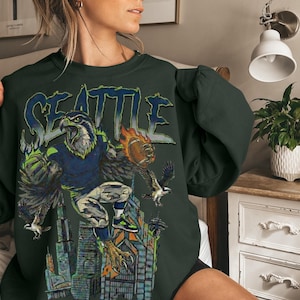 May include: Dark green sweatshirt featuring a graphic of a Seattle Seahawks eagle with a football, the word "SEATTLE" in blue, and a cityscape. The design includes shades of blue, green, orange, and gray.