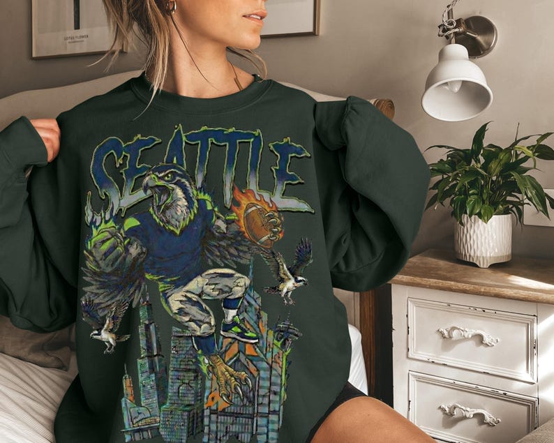 May include: Dark green sweatshirt featuring a graphic of a Seattle Seahawks eagle with a football, the word "SEATTLE" in blue, and a cityscape. The design includes shades of blue, green, orange, and gray.