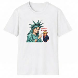 May include: White t-shirt featuring a cartoon illustration of the Statue of Liberty pointing at a pig wearing a suit. The text "QUIET PIGGY" is printed in red. The Statue of Liberty is depicted with teal skin and a stern expression.