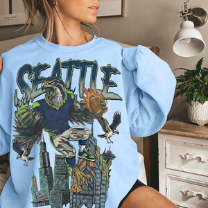 May include: Light blue sweatshirt featuring a graphic of a hawk in a football uniform, with the word "SEATTLE" above. The hawk is holding a football and is set against a cityscape background. The design incorporates green, blue, and orange.