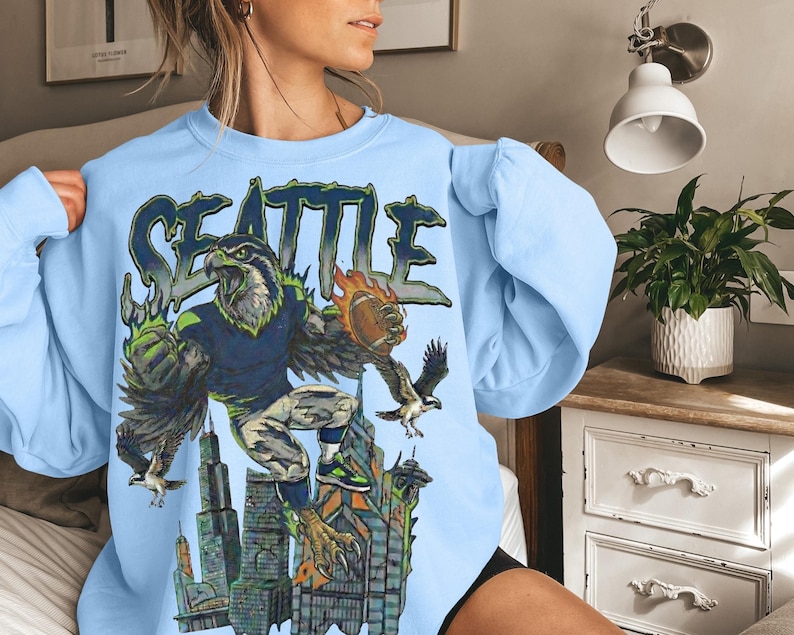 May include: Light blue sweatshirt featuring a graphic of a hawk in a football uniform, with the word "SEATTLE" above. The hawk is holding a football and is set against a cityscape background. The design incorporates green, blue, and orange.