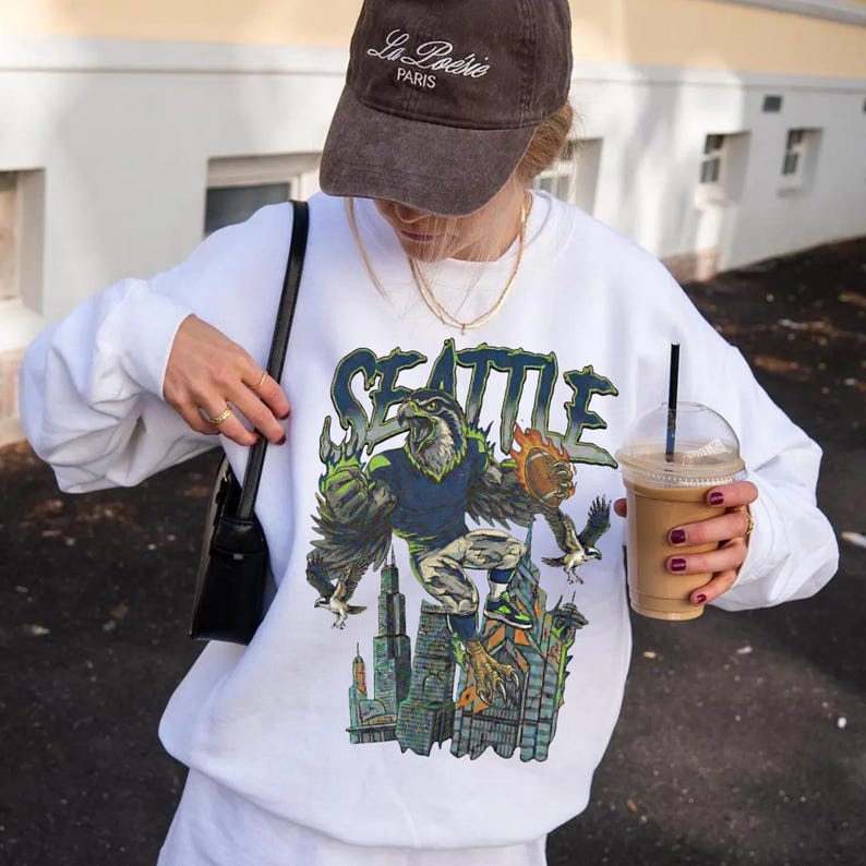 May include: White long-sleeve sweatshirt with a graphic of a Seattle-themed eagle character over a cityscape. The word "SEATTLE" is above the graphic in a stylized font. The person is wearing a brown cap with "La Po&eacute;sie PARIS" on it.