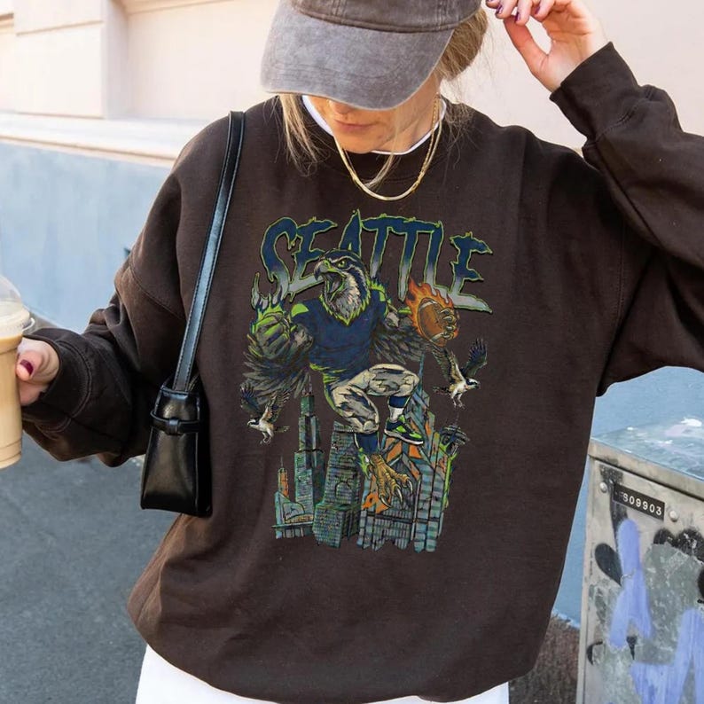 May include: A brown sweatshirt with a graphic featuring the word "SEATTLE" in blue and green, along with an eagle-like figure holding a football. The graphic is set against a cityscape.