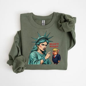 May include: Olive green sweatshirt featuring a graphic of the Statue of Liberty pointing at a pig in a suit, with the text "Quiet Piggy". The Statue of Liberty is depicted with teal hair and a stern expression.