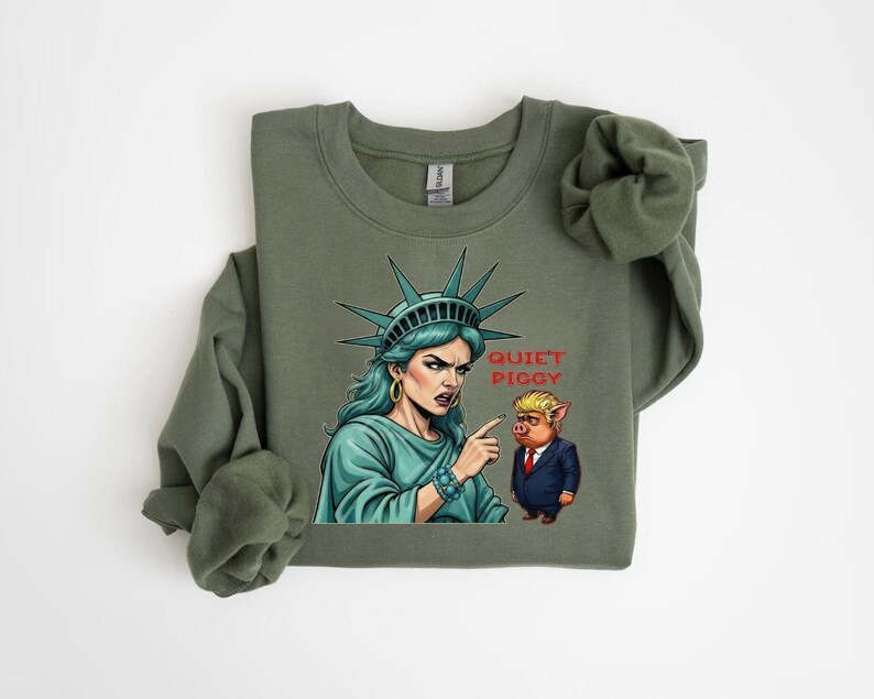 May include: Olive green sweatshirt featuring a graphic of the Statue of Liberty pointing at a pig in a suit, with the text "Quiet Piggy". The Statue of Liberty is depicted with teal hair and a stern expression.