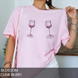 May include: A light pink short-sleeve t-shirt featuring two dark purple wine glass illustrations. The left glass is labeled "The Bride" and the right glass is labeled "Emma's Bachelorette Napa Valley 2024". The shirt has a crew neckline.