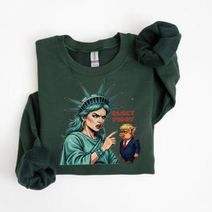 May include: Dark green sweatshirt featuring a graphic of the Statue of Liberty pointing at a pig wearing a suit. The text "Quiet Piggy" is printed on the sweatshirt. The Statue of Liberty is depicted with teal skin and a stern expression.
