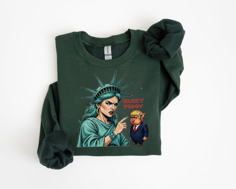 May include: Dark green sweatshirt featuring a graphic of the Statue of Liberty pointing at a pig wearing a suit. The text "Quiet Piggy" is printed on the sweatshirt. The Statue of Liberty is depicted with teal skin and a stern expression.