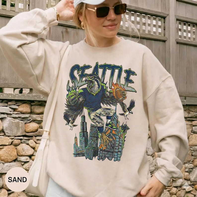 May include: Sand-colored sweatshirt featuring a graphic of a Seattle-themed eagle with a basketball and city skyline. The word "SEATTLE" is printed above the eagle in blue and green. The sweatshirt has long sleeves and a crew neck.