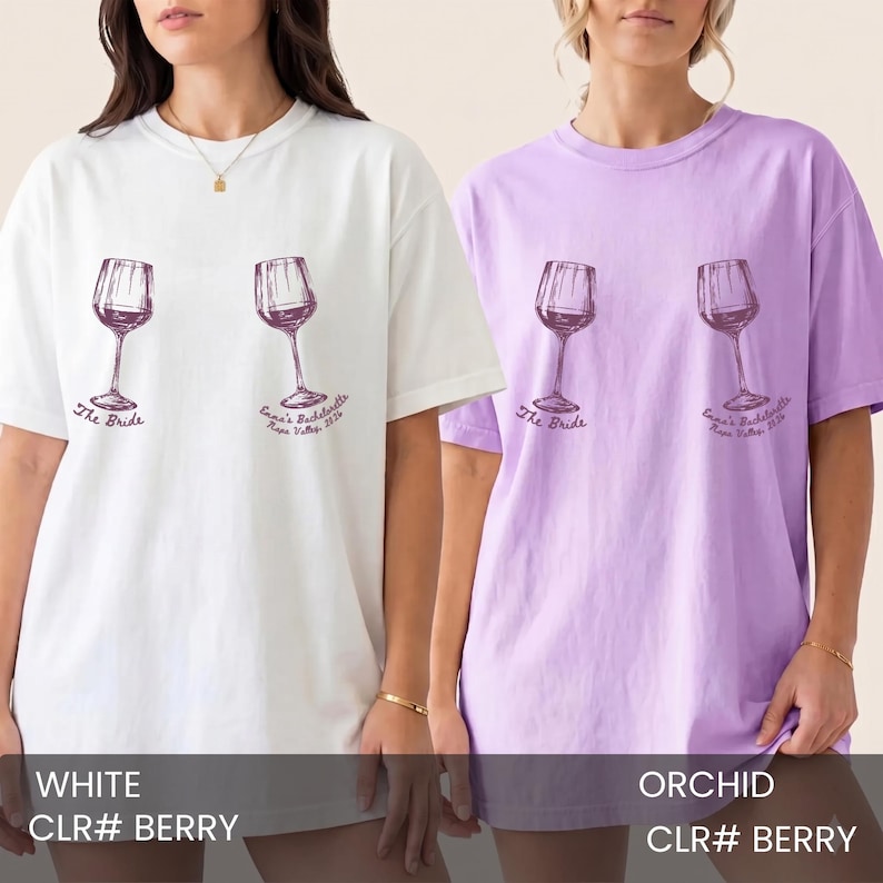 May include: Two short-sleeve crew neck t-shirts, one white and one orchid, each featuring a dark purple graphic of two wine glasses. Below the glasses, the text reads The Bride and Emma's Bachelorette Napa Valley 2024 in a cursive script.
