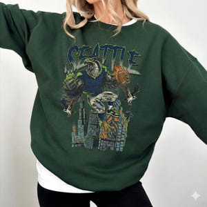 May include: Dark green sweatshirt featuring a graphic of a football-playing eagle with the word "SEATTLE" above. The eagle is depicted in a football uniform, holding a football, and standing on a cityscape. The sweatshirt has a crew neck and long sleeves.