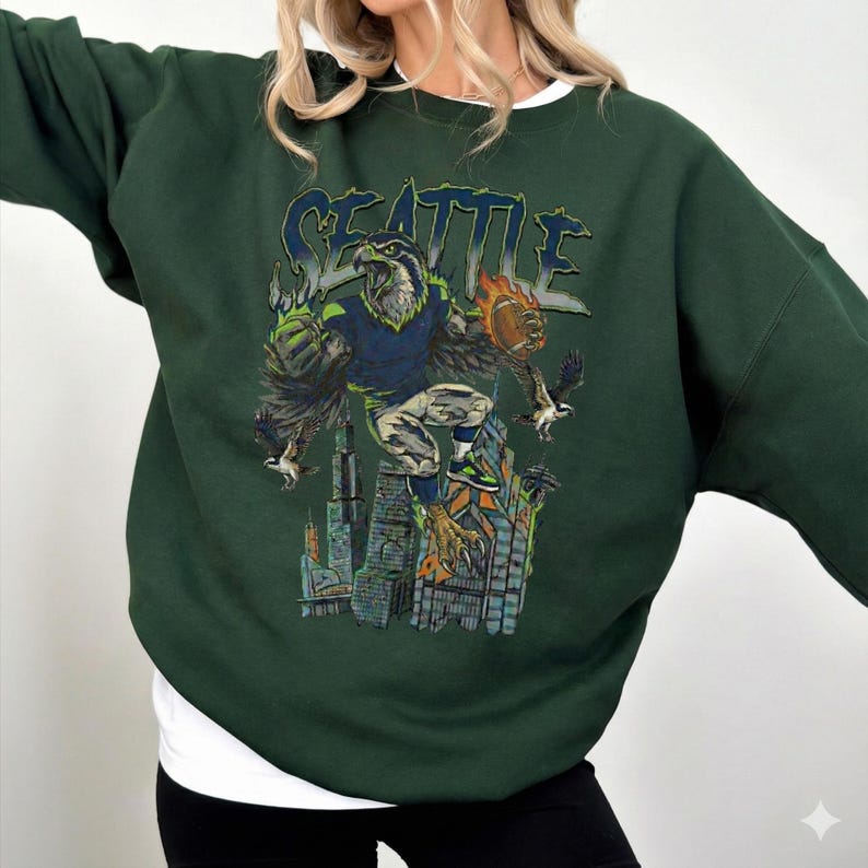 May include: Dark green sweatshirt featuring a graphic of a football-playing eagle with the word "SEATTLE" above. The eagle is depicted in a football uniform, holding a football, and standing on a cityscape. The sweatshirt has a crew neck and long sleeves.
