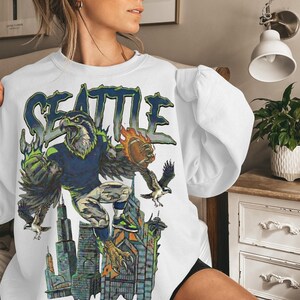 May include: White sweatshirt featuring a graphic of a football-playing eagle with the word "SEATTLE" in blue and green. The eagle is depicted with a football and is set against a cityscape background.