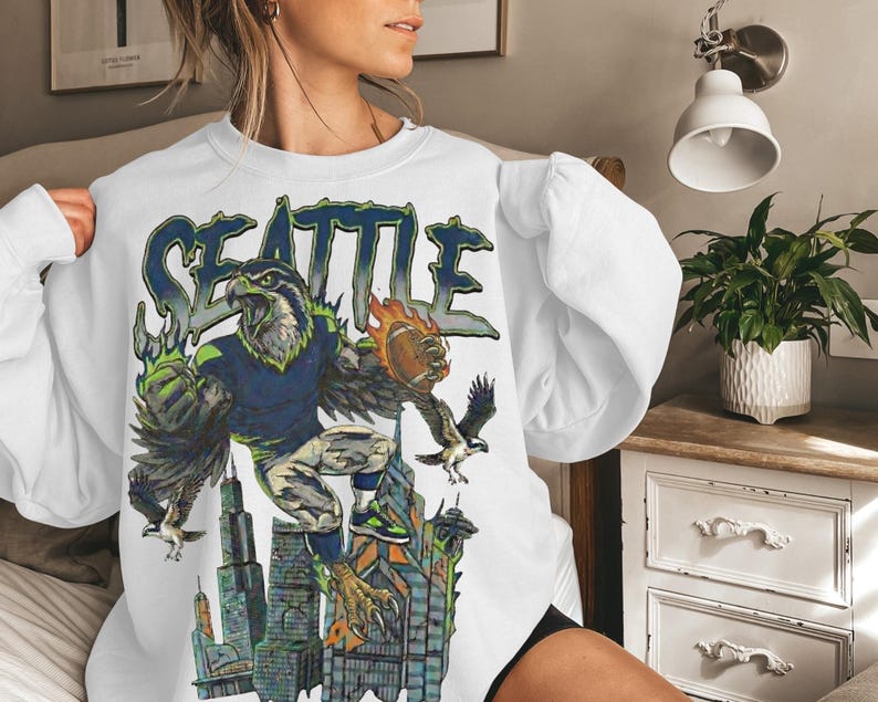 May include: White sweatshirt featuring a graphic of a football-playing eagle with the word "SEATTLE" in blue and green. The eagle is depicted with a football and is set against a cityscape background.
