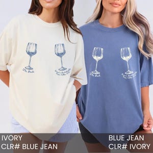 May include: Two short-sleeve crewneck t-shirts, one ivory and one blue jean, each featuring a graphic of two wine glasses. The ivory shirt has blue text reading The Bride and Emma's Bachelorette Napa Valley 2024. The blue shirt has white text.
