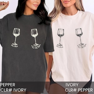 May include: A charcoal gray t-shirt and an ivory t-shirt, each featuring two wine glass illustrations. The charcoal shirt displays the text The Bach and Emma's Bachelorette Napa Valley 2026. The ivory shirt displays The Bride and Emma's Bachelorette Napa Valley 2026.