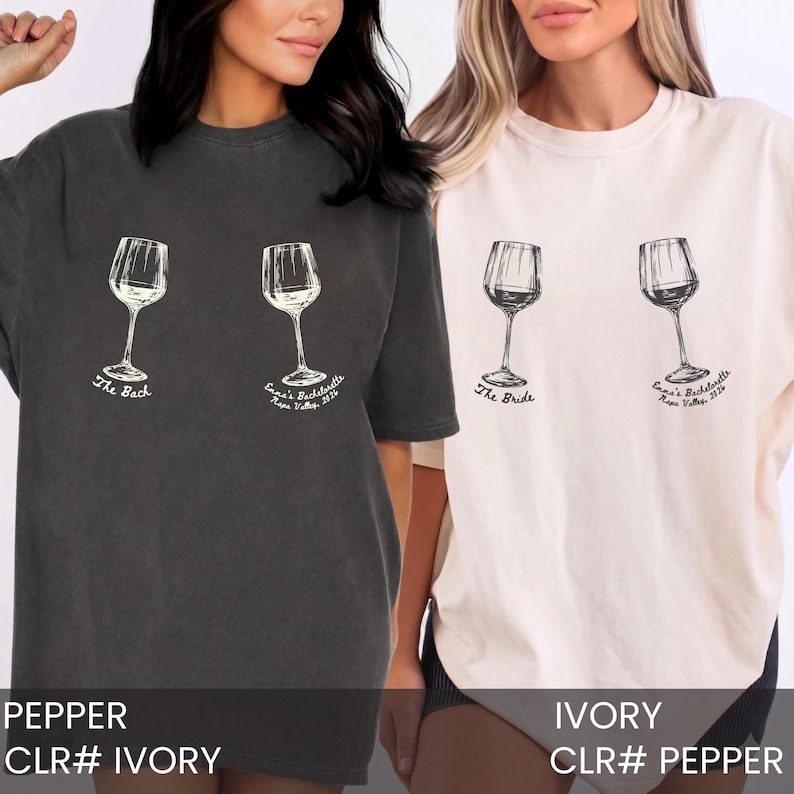 May include: A charcoal gray t-shirt and an ivory t-shirt, each featuring two wine glass illustrations. The charcoal shirt displays the text The Bach and Emma's Bachelorette Napa Valley 2026. The ivory shirt displays The Bride and Emma's Bachelorette Napa Valley 2026.