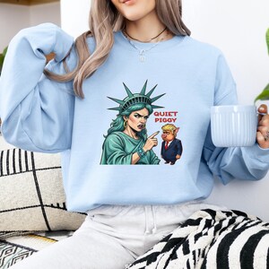 May include: Light blue sweatshirt featuring a graphic of the Statue of Liberty pointing at a cartoon pig with the text "QUIET PIGGY". The Statue of Liberty is depicted with green skin and a stern expression. The pig is wearing a suit.
