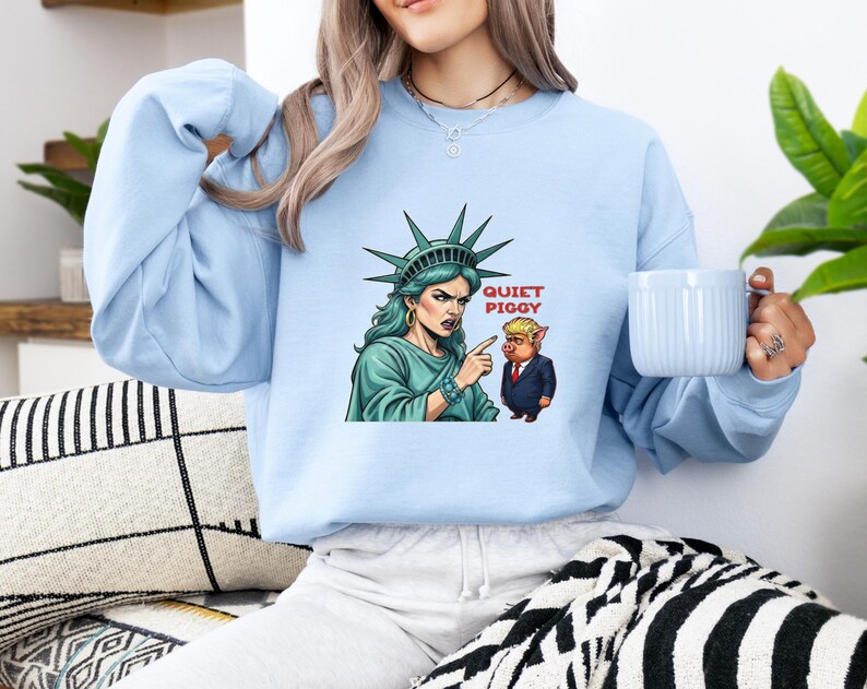 May include: Light blue sweatshirt featuring a graphic of the Statue of Liberty pointing at a cartoon pig with the text "QUIET PIGGY". The Statue of Liberty is depicted with green skin and a stern expression. The pig is wearing a suit.