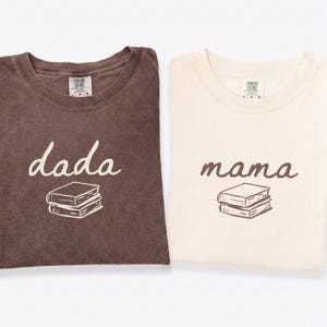 Może przedstawiać: Dwie koszulki Comfort Colors, jedna brązowa i jedna kremowa. Brązowa koszulka ma nadruk z napisem "dada" i stos książek. Kremowa koszulka ma nadruk z napisem "mama" i stos książek.