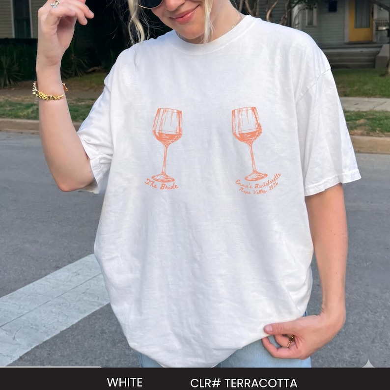 May include: A white short-sleeved t-shirt featuring two orange line-art illustrations of wine glasses. The left glass has the text "The Bride" underneath, and the right glass has the text "Emma's Bachelorette Napa Valley, 2024" printed below it.