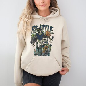 May include: Beige hoodie with a graphic of a Seattle-themed eagle character holding a football, with the city skyline in the background. The word "SEATTLE" is arched above the eagle in a green and blue font. The hoodie has a kangaroo pocket and a drawstring hood.