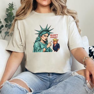 May include: Beige t-shirt featuring a graphic of the Statue of Liberty pointing at a pig in a suit, with the text "QUIET PIGGY" in red. The Statue of Liberty is depicted in teal, and the pig is wearing a red tie.