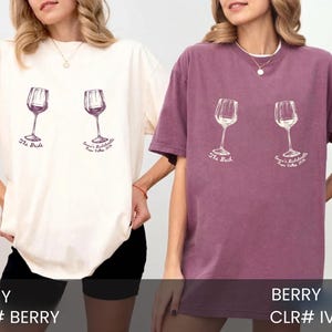 May include: Two short-sleeve crewneck t-shirts, one ivory and one berry, each featuring a graphic of two wine glasses. The ivory shirt has purple text reading The Bride and Emma's Bachelorette Napa Valley 2024. The berry shirt has white text.