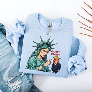 May include: Light blue sweatshirt featuring a graphic of the Statue of Liberty pointing at a cartoon pig with the text "QUIET PIGGY". The sweatshirt is folded next to a pair of blue jeans.