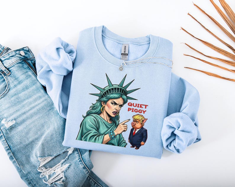 May include: Light blue sweatshirt featuring a graphic of the Statue of Liberty pointing at a cartoon pig with the text "QUIET PIGGY". The sweatshirt is folded next to a pair of blue jeans.