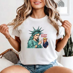 May include: White t-shirt featuring a graphic of the Statue of Liberty pointing at a pig wearing a suit. The text "QUIET PIGGY" is printed in red. The Statue of Liberty is depicted with blue hair and a green robe.