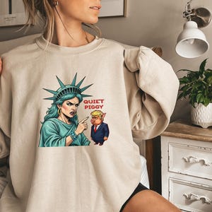 May include: Beige sweatshirt featuring a graphic of the Statue of Liberty pointing at a pig in a suit, with the text "QUIET PIGGY". The Statue of Liberty is depicted with teal hair and a stern expression. The pig is wearing a suit and tie.