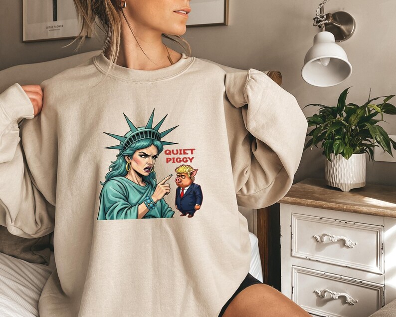 May include: Beige sweatshirt featuring a graphic of the Statue of Liberty pointing at a pig in a suit, with the text "QUIET PIGGY". The Statue of Liberty is depicted with teal hair and a stern expression. The pig is wearing a suit and tie.