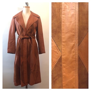 May include: A long, brown leather trench coat with a matching belt. The coat is displayed on a mannequin. The right side of the image shows a close-up of the leather and suede patchwork design.