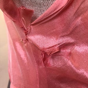 May include: Close-up of a pink, shiny fabric with a V-neck design. The fabric appears to be slightly damaged, with some fraying at the seams. The material has a subtle sheen, and the color is a soft, light pink.