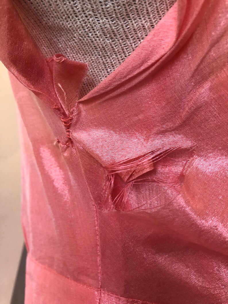 May include: Close-up of a pink, shiny fabric with a V-neck design. The fabric appears to be slightly damaged, with some fraying at the seams. The material has a subtle sheen, and the color is a soft, light pink.