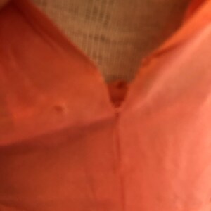 May include: Close-up of a coral-colored garment with a V-neckline. The fabric appears smooth and possibly lightweight, with a button detail at the neckline. The image focuses on the upper portion of the garment.