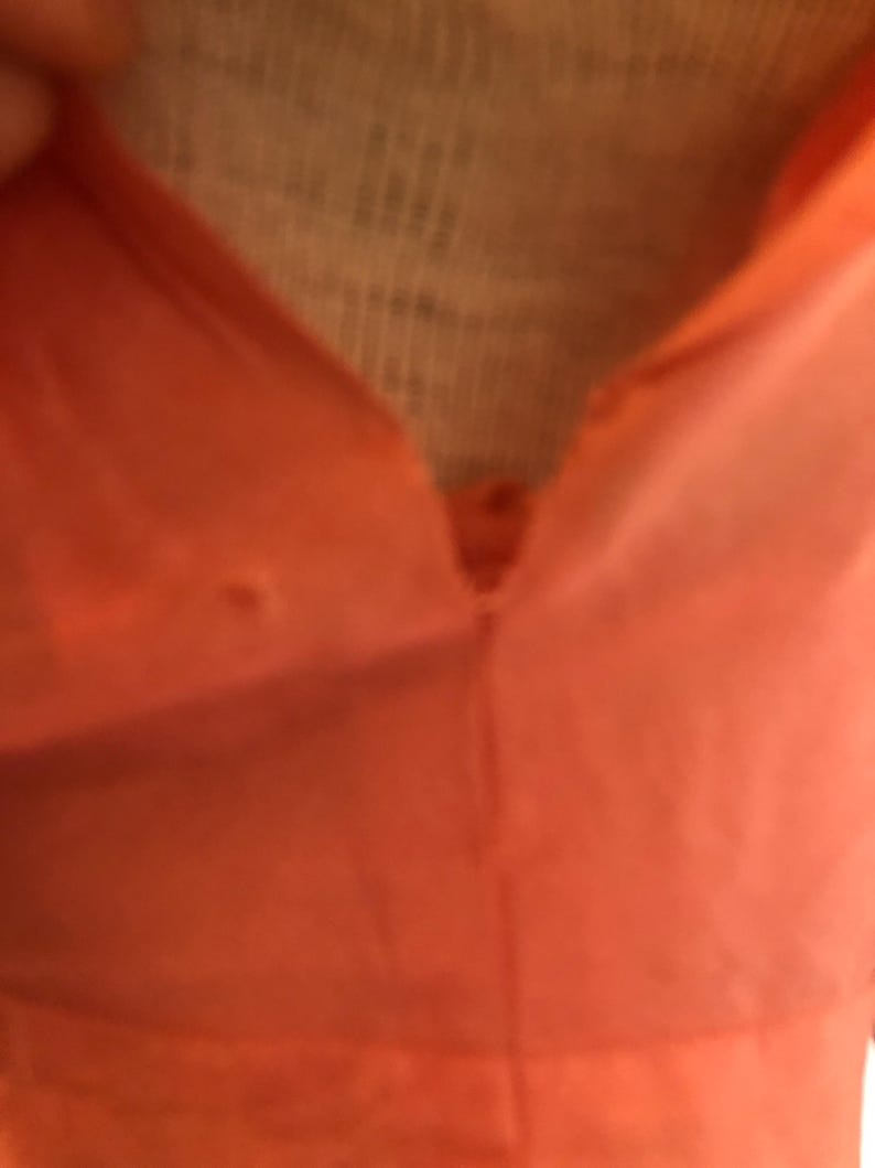 May include: Close-up of a coral-colored garment with a V-neckline. The fabric appears smooth and possibly lightweight, with a button detail at the neckline. The image focuses on the upper portion of the garment.
