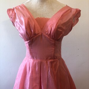 May include: A vintage pink dress with a V-neckline and short, puffed sleeves. The dress features a fitted bodice and a flared skirt, with pleats at the waist. The fabric has a subtle sheen, giving it a delicate appearance. The dress is displayed on a mannequin.