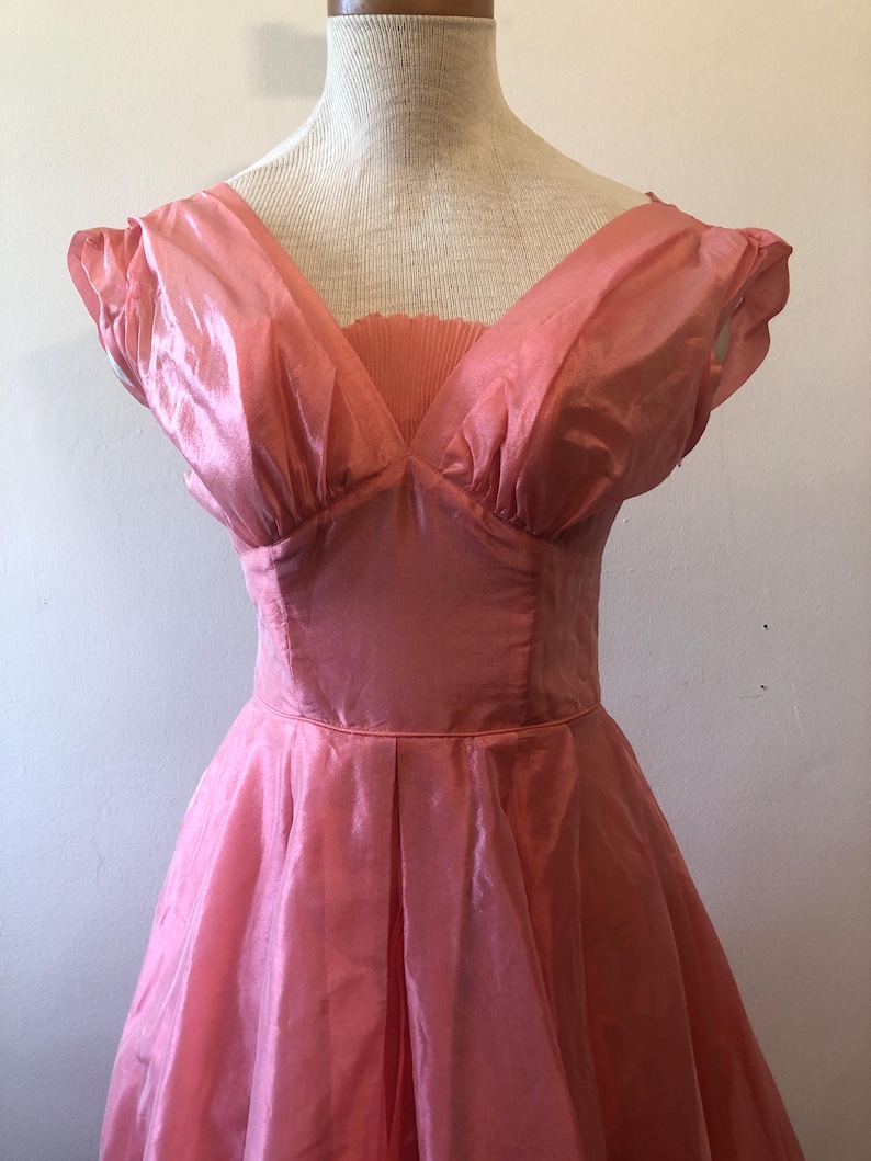 May include: A vintage pink dress with a V-neckline and short, puffed sleeves. The dress features a fitted bodice and a flared skirt, with pleats at the waist. The fabric has a subtle sheen, giving it a delicate appearance. The dress is displayed on a mannequin.