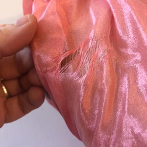May include: Close-up of a section of a pink fabric, possibly silk or a similar material. The fabric has a shiny, reflective quality. There is a tear in the fabric, with frayed edges visible. The fabric is being held by a hand.